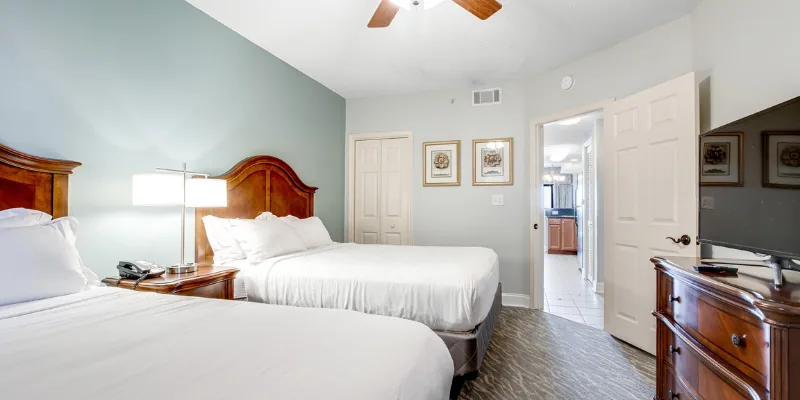 A wide view of a hotel room with two double beds, a ceiling fan, a flat-screen TV on a dresser, and a doorway leading to another room.