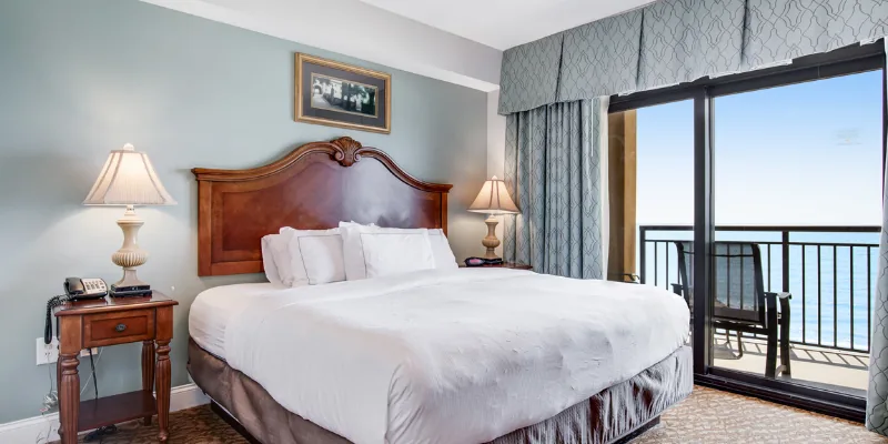 A wide-angle view of a hotel room with a large bed, two nightstands, and a balcony overlooking the ocean.