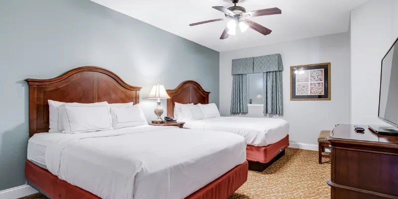 A hotel room with two beds, a ceiling fan, a window with curtains, a flat-screen TV on a dresser, and a lamp on a nightstand.