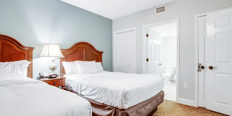 A hotel room with two twin beds, wooden headboards, and a nightstand with a lamp between them, with a doorway to the bathroom visible.