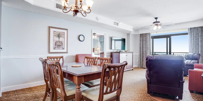 A wide view of a dining area with a table, chairs, and a sofa, with a balcony door and a view outside.