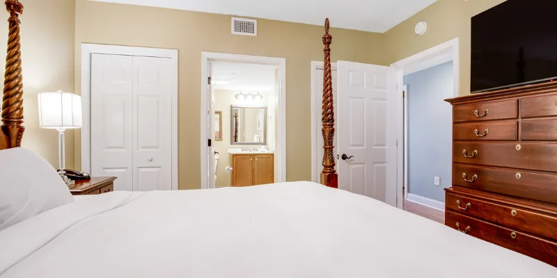 A wide view of a hotel room with a four-poster bed, wooden dresser, and flat-screen TV on top, with a bathroom and closet in the background.