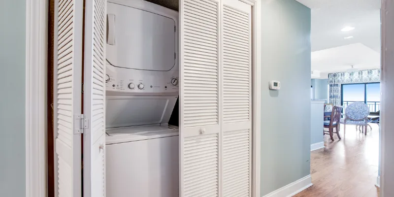 A laundry closet with a stacked washer and dryer, white closet doors, and a view into a dining area.
