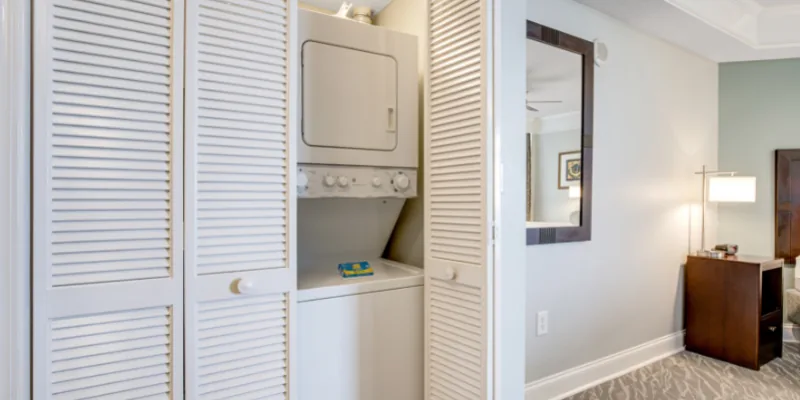 A laundry area with a washer and dryer, closet doors, a mirror, lamp, and cabinet in a room.