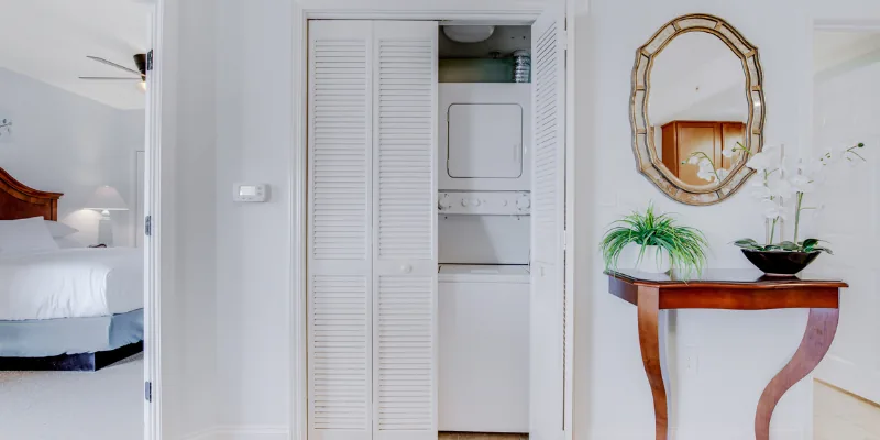 A laundry closet with a washer and dryer, a console table with plants and a mirror, and a view of a bed in the bedroom.