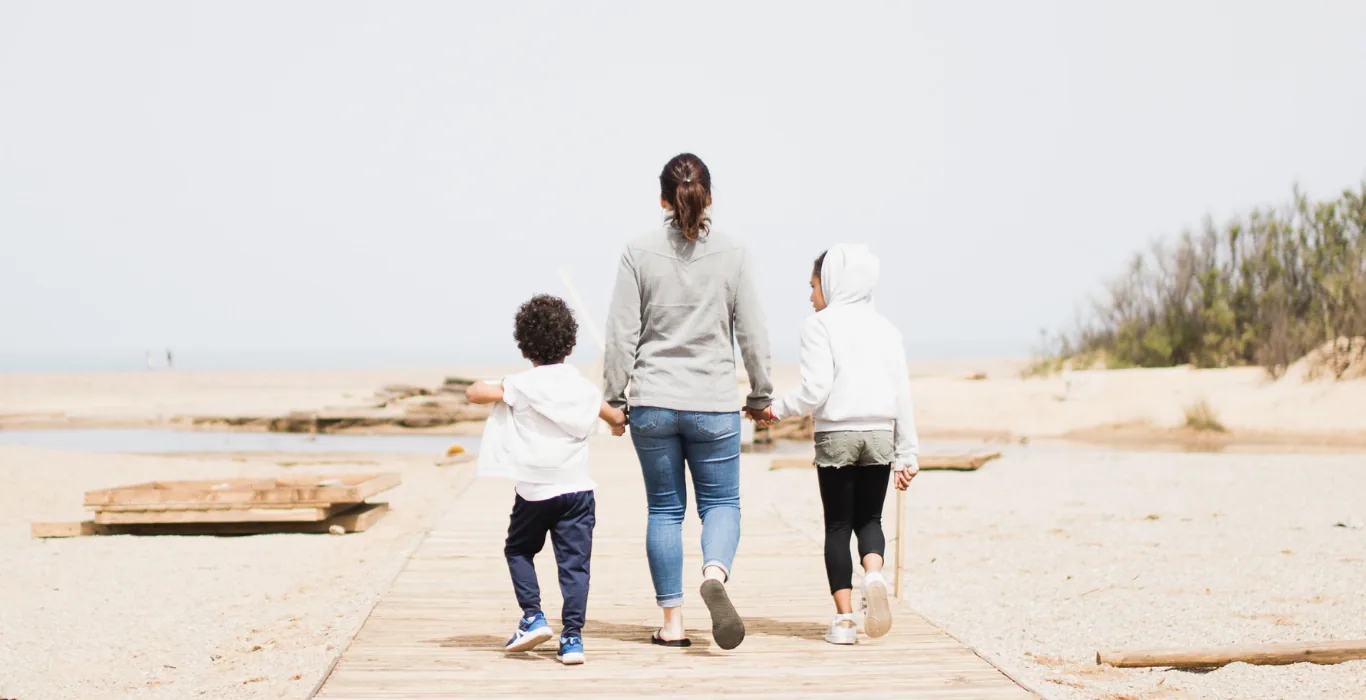A woman and two children walking on a boardwalk towards the beach.